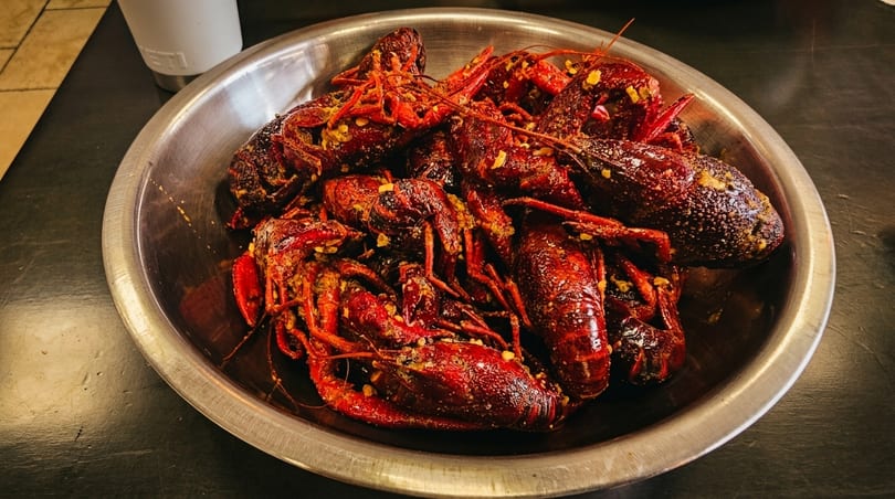 A large metal bowl filled with bright red, seasoned crawfish on a dark table.